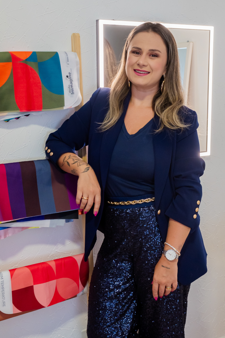 Juliana Tait sorrindo, segurando um cabide com uma blusa cinza detalhada com pedras, em seu ateliê de curadoria